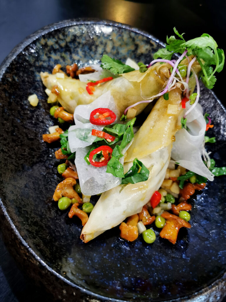 Knusprige Frühlingsrollen mit Erbsen und Gemüse, garniert mit Chili, frischen Kräutern und feinen Gemüsestreifen in einer schwarzen Schale.