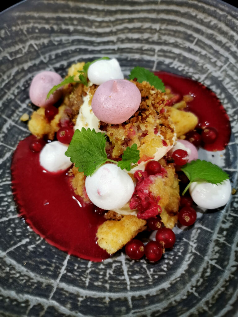 Dessert mit Vanillecreme, Beerenkompott, roter Fruchtsauce, knusprigem Crumble und frischen Johannisbeeren, garniert mit Minzblättern.