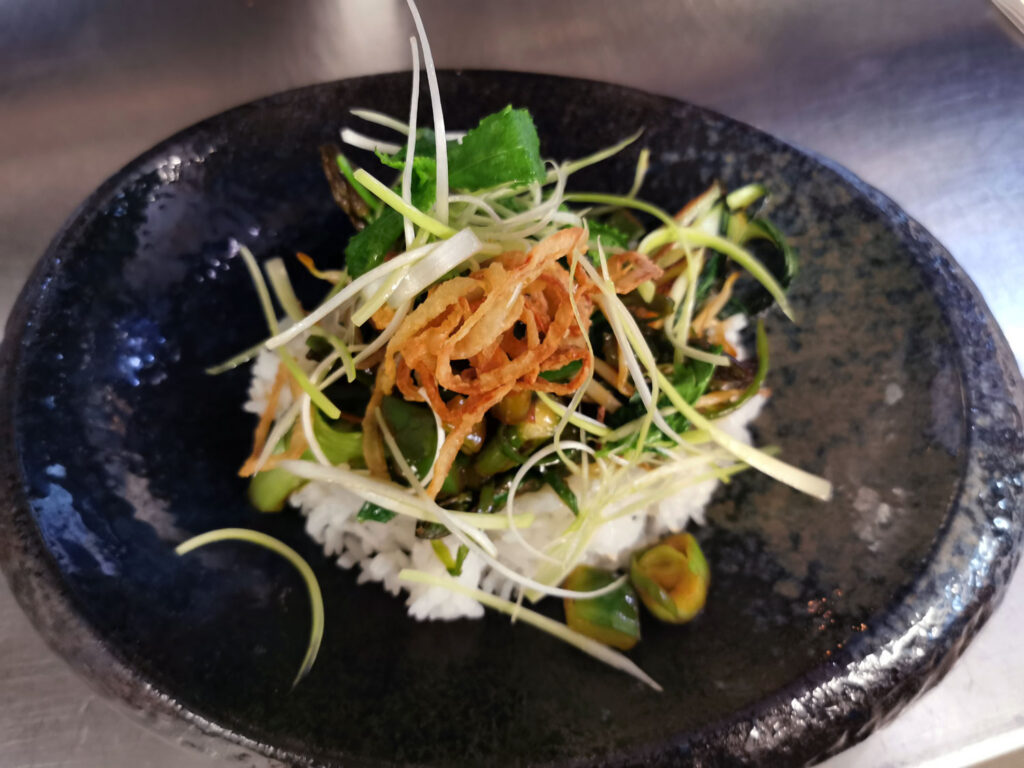 Kreativ angerichtetes Reisgericht mit grünem Spargel, frischen Kräutern und feinen Gemüsestreifen, serviert in einer schwarzen Schale.
