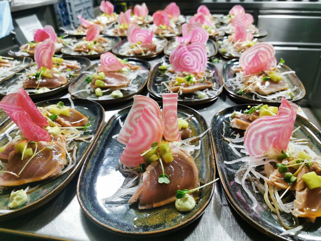 Mehrere kunstvoll angerichtete Vorspeisen mit Fisch, eingelegter Ringelbete, Avocadowürfeln und Sprossen stehen servierbereit auf schwarzen Tellern in einer professionellen Küche.