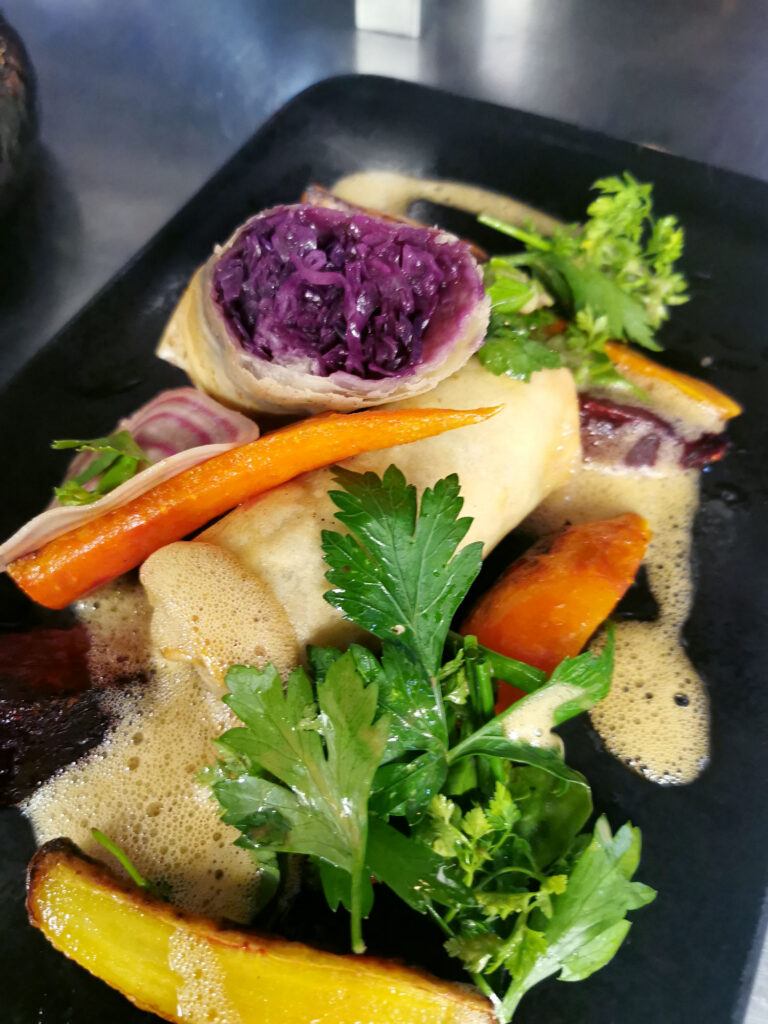 Kreativ angerichtetes Gourmetgericht mit knusprigen Rollen, Rotkraut, glasierter Karotte, frischen Kräutern und einer luftigen Sauce auf einem schwarzen Teller.
