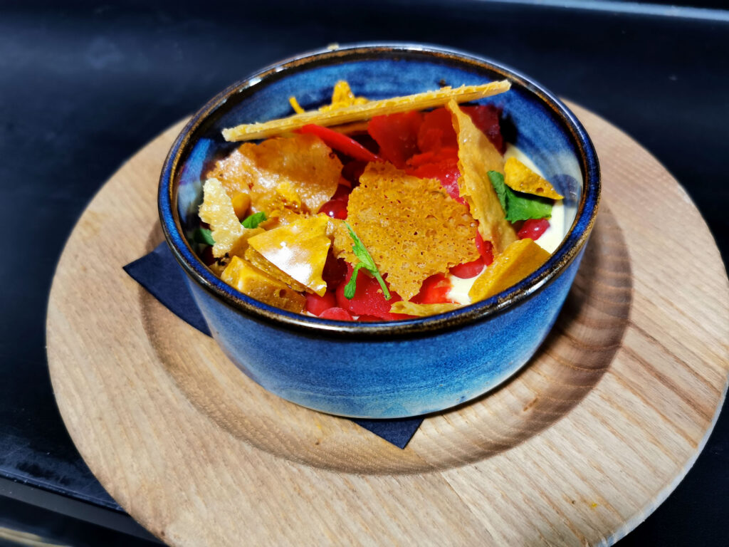 Modern angerichtetes Dessert in einer blauen Schale mit cremiger Basis, fruchtigen Komponenten und frischen Kräutern, serviert auf einem Holzbrett.
