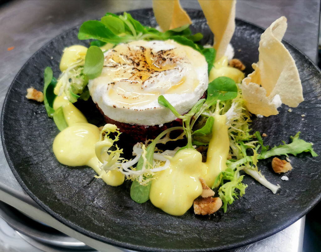 Rund angerichtete Speise mit Blattsalat, cremiger Sauce, Walnüssen und knusprigen Chips auf einem schwarzen Teller.