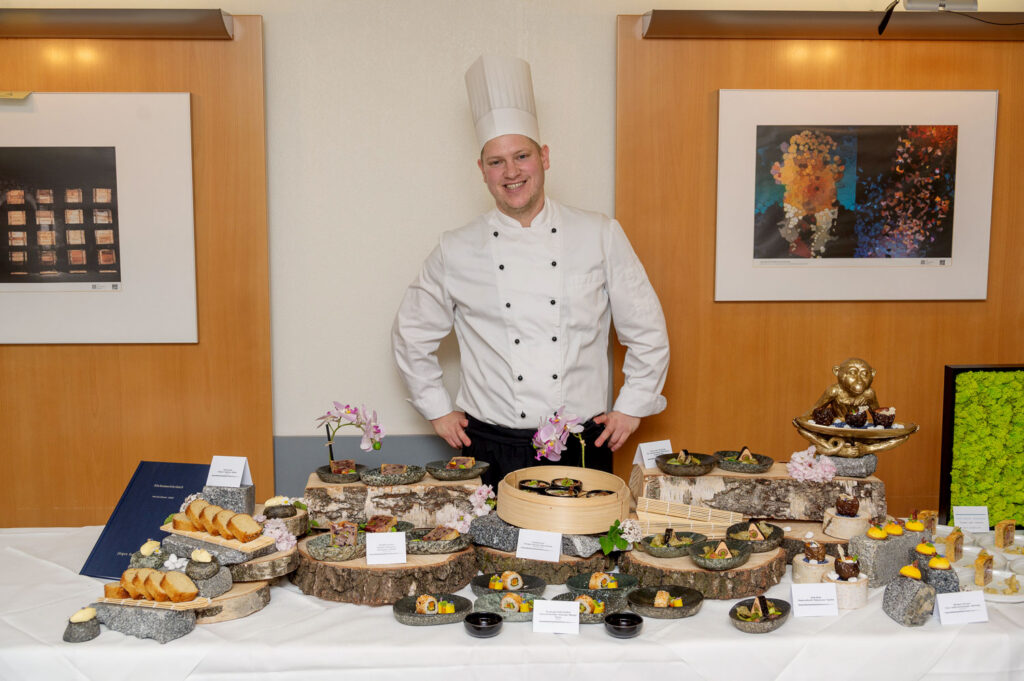 Koch in weißer Kochjacke und Kochmütze präsentiert ein Buffet mit kunstvoll angerichteten Vorspeisen und Fingerfood.