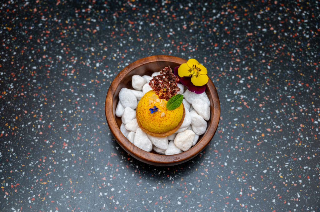 Kunstvoll angerichtetes Dessert mit einer gelben Mousse-Tartelette, essbaren Blüten, Minze und knusprigem Dekor, serviert in einer Holzschale auf weißen Kieselsteinen.