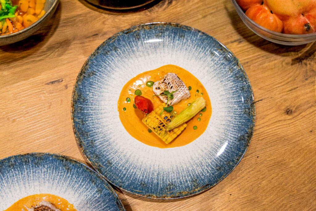 Kunstvoll angerichteter Hauptgang mit gebratenem Fisch, Mais, Babymais und Tomate auf einer cremigen orangefarbenen Sauce, serviert auf einem großen Keramikteller.