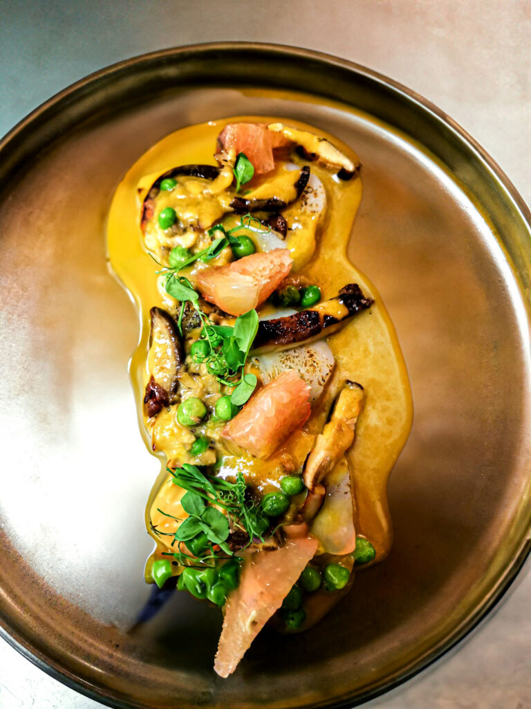 Kreativ angerichtetes Gourmetgericht mit gebratenen Jakobsmuscheln, Pilzen, Erbsen und cremiger Sauce, garniert mit frischen Kräutern auf einem eleganten Teller.